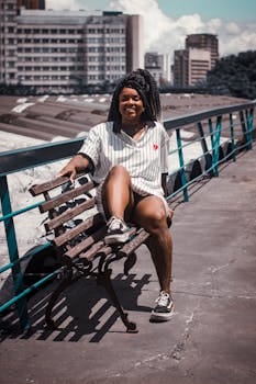 A woman with dreadlocks sitting confidently on a bench in an urban rooftop setting, enjoying the sunny day.
